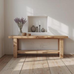 Può includere: Una consolle in legno chiaro con un bordo naturale. Il tavolo ha un ripiano inferiore e gambe spesse. Un vaso di vetro con lavanda, due ciotole intrecciate e oggetti decorativi sono sul tavolo. Lo sfondo è un muro bianco con una mensola incassata.