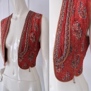 Puede incluir: Un chaleco acolchado de color rojo anaranjado, adornado con un estampado de cachemira en tonos azules, marrones y crema. El chaleco presenta un ribete marrón y un patrón decorativo en zigzag en los bordes. Pequeños dijes metálicos cuelgan de la parte inferior.