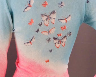 Könnte beinhalten: Ein hellblau-rosa Pullover mit Schmetterlingsmuster. Der Pullover hat einen Farbverlauf, der von Hellblau oben nach Rosa unten verläuft. Der Rücken des Pullovers ist mit Schmetterlingen in verschiedenen Größen verziert.