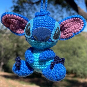 May include: A handmade crochet figure of a blue alien character with large ears, black eyes, and a dark blue nose. The figure is made with blue and light blue yarn, with pink accents on the ears. It is a whimsical, handcrafted item.