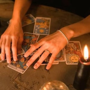 Può includere: Un set di carte dei tarocchi sparse su una superficie con una candela nera accesa nelle vicinanze. Le carte presentano varie illustrazioni, tra cui un simbolo solare e motivi circolari. Sono visibili un sottile braccialetto metallico e due semplici anelli.
