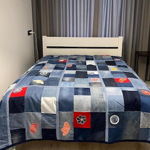 May include: A patchwork quilt made from denim squares in various shades of blue, with red, white, and floral accents. The quilt is spread across a white-framed bed, creating a cozy and handcrafted aesthetic. The quilt's design is a mix of solid and patterned denim.