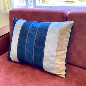 May include: A rectangular pillow with a striped design in shades of blue, white, and beige. The pillow is placed on a red sofa. The pillow's design features vertical stripes, creating a modern and stylish look. The pillow is a decorative item.