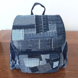 May include: A denim backpack with a patchwork design. The backpack features a top handle, a front pocket, and side pockets. The denim is a mix of light and dark blue patches, creating a unique, textured look. Zippers are visible on the pockets.