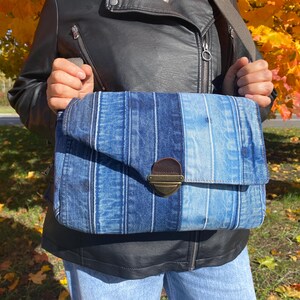 K&ouml;nnte beinhalten: Eine blaue Jeans-Umh&auml;ngetasche mit Klappenverschluss und messingfarbenem Verschluss. Die Tasche hat vertikale Streifen in verschiedenen Blaut&ouml;nen. Die Tasche wird mit einem braunen Riemen &uuml;ber der Schulter getragen.