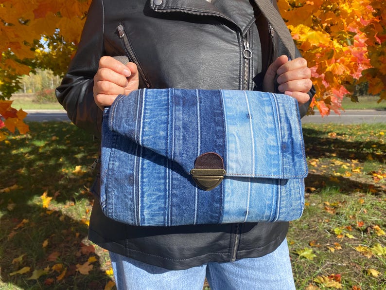 K&ouml;nnte beinhalten: Eine blaue Jeans-Umh&auml;ngetasche mit Klappenverschluss und messingfarbenem Verschluss. Die Tasche hat vertikale Streifen in verschiedenen Blaut&ouml;nen. Die Tasche wird mit einem braunen Riemen &uuml;ber der Schulter getragen.