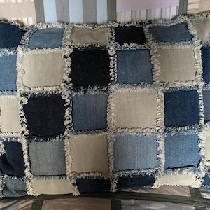 Puede incluir: Cojín rectangular con diseño de patchwork. El cojín presenta cuadrados de varios tonos de mezclilla, incluyendo azul claro, medio y oscuro, y algunos cuadrados de color beige claro. Los bordes de cada cuadrado están deshilachados, agregando textura.