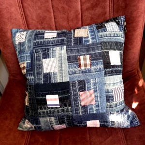 Può includere: Cuscino quadrato in denim con un design patchwork. Il cuscino presenta varie tonalità di quadrati di denim blu, accentuati da piccole toppe di tessuto in bianco, rosso e tessuti fantasia. Il cuscino è appoggiato su una sedia rossa.
