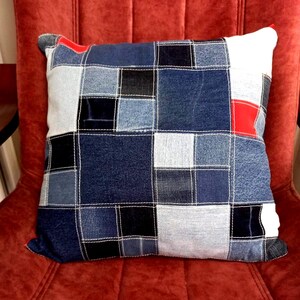 Puede incluir: Cojín cuadrado de patchwork de mezclilla con cuadrados de mezclilla de varios tonos de azul, negro y blanco. El cojín tiene un acento rojo y costuras blancas. El cojín está sobre una silla roja.