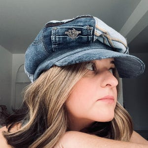Puede incluir: Gorra de estilo newsboy de mezclilla con un pequeño bolsillo y un parche que dice "FASHION JEANS". La gorra es un patchwork de mezclilla y tela a cuadros. Tiene una pequeña visera y una corona redondeada.