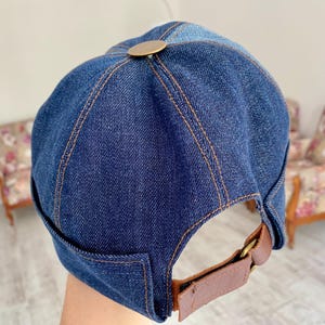 May include: A blue denim baseball cap with a brown leather strap and a brass button on top.