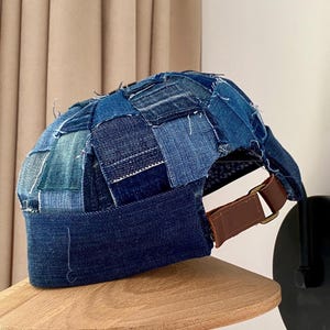Puede incluir: Una gorra de béisbol de mezclilla azul con parches, con una correa de cuero marrón y una hebilla de latón.