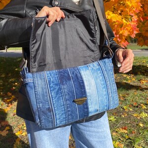 K&ouml;nnte beinhalten: Rechteckige Jeans-Umh&auml;ngetasche mit einem Farbverlauf in Blaut&ouml;nen. Die Tasche hat einen Klappenverschluss mit einem bronzefarbenen Verschluss und ein schwarzes Innenfutter. Die Tasche wird mit einer schwarzen Jacke und einer blauen Jeans getragen.