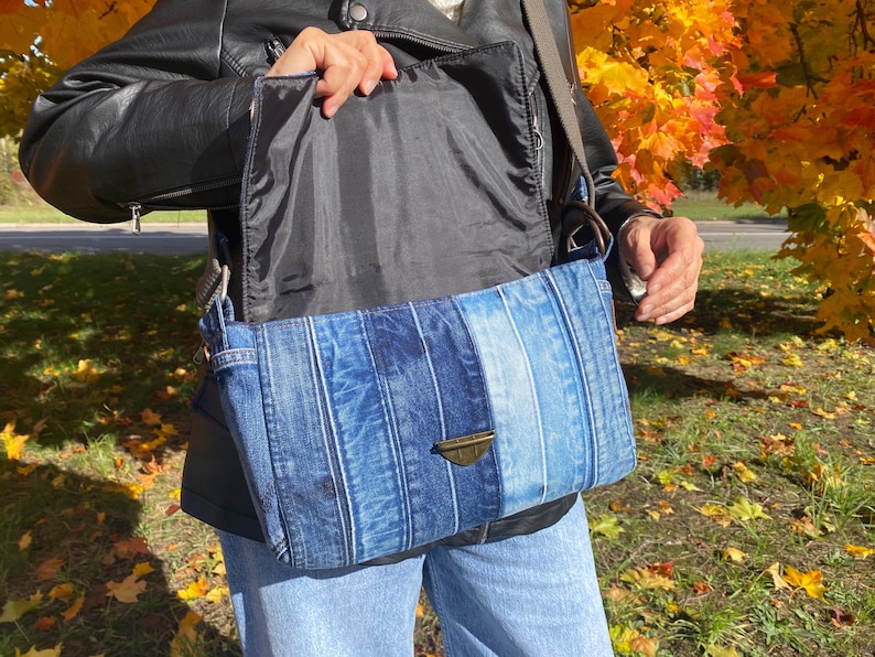 K&ouml;nnte beinhalten: Rechteckige Jeans-Umh&auml;ngetasche mit einem Farbverlauf in Blaut&ouml;nen. Die Tasche hat einen Klappenverschluss mit einem bronzefarbenen Verschluss und ein schwarzes Innenfutter. Die Tasche wird mit einer schwarzen Jacke und einer blauen Jeans getragen.