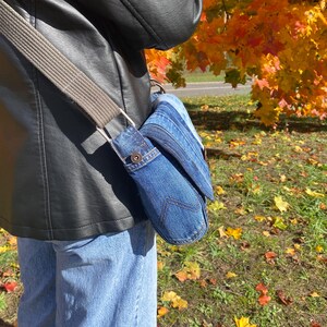 K&ouml;nnte beinhalten: Eine Jeans-Umh&auml;ngetasche mit grauem Riemen wird &uuml;ber der Schulter getragen. Die Tasche besteht aus blauem Jeansstoff und hat einen Klappenverschluss. Der Hintergrund zeigt Herbstlaub mit gelben und orangen Bl&auml;ttern.