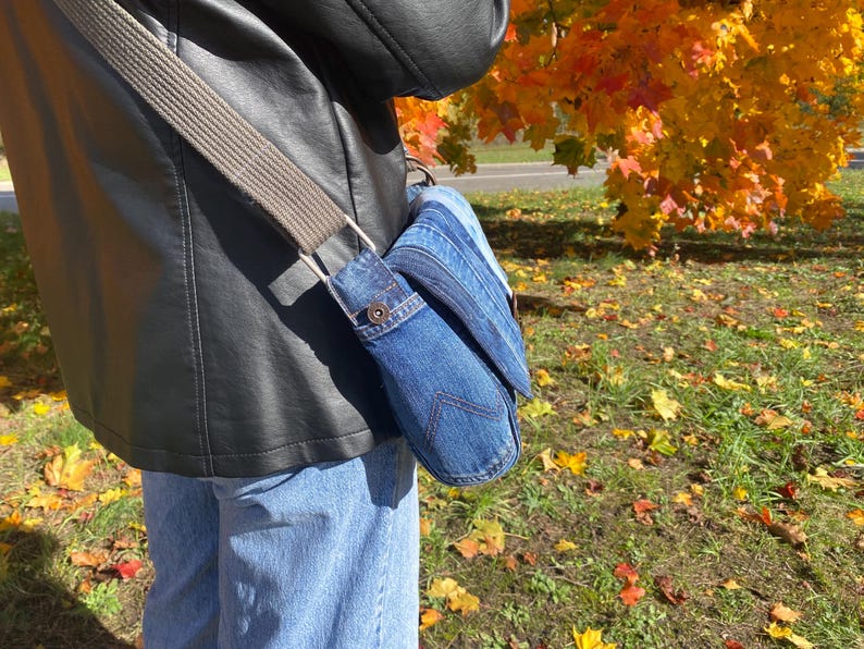 K&ouml;nnte beinhalten: Eine Jeans-Umh&auml;ngetasche mit grauem Riemen wird &uuml;ber der Schulter getragen. Die Tasche besteht aus blauem Jeansstoff und hat einen Klappenverschluss. Der Hintergrund zeigt Herbstlaub mit gelben und orangen Bl&auml;ttern.