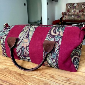 May include: A burgundy duffel bag with a floral tapestry design. The bag features a central burgundy panel flanked by tapestry panels with brown leather handles and accents. The bag is on a wooden surface.