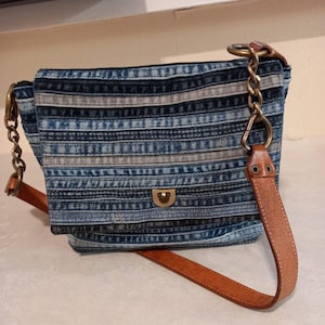 Może przedstawiać: Torebka listonoszka z denimu w niebiesko-białe paski, z brązowym paskiem skórzanym i złotym łańcuszkiem. Torebka ma klapkę zamykaną na złotą klamrę.