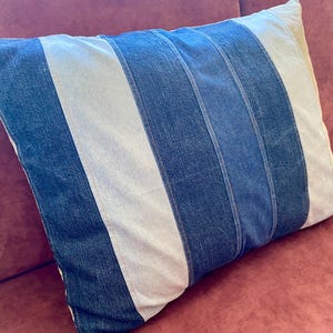 Puede incluir: Cojín rectangular con diseño de mezclilla. El cojín presenta rayas verticales en tonos azules y blancos. El cojín está colocado sobre una superficie de tonos rojizos.