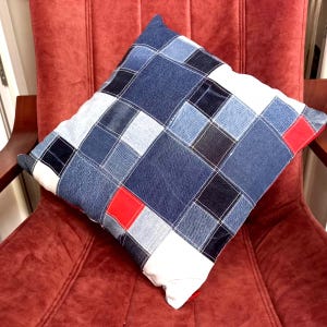 May include: Square denim patchwork pillow with a variety of blue shades, white stitching, and red accent squares. The pillow is placed on a red chair, showcasing its design and texture.