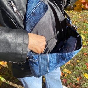 K&ouml;nnte beinhalten: Eine blaue Jeans-Umh&auml;ngetasche mit Patchwork-Design. Die Tasche hat ein schwarzes Innenfutter und einen schwarzen Riemen. Die Person tr&auml;gt eine schwarze Lederjacke und blaue Jeans. Die Tasche ist offen und zeigt einen dunkelblauen Gegenstand im Inneren.