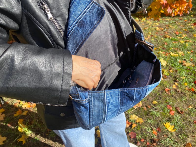 K&ouml;nnte beinhalten: Eine blaue Jeans-Umh&auml;ngetasche mit Patchwork-Design. Die Tasche hat ein schwarzes Innenfutter und einen schwarzen Riemen. Die Person tr&auml;gt eine schwarze Lederjacke und blaue Jeans. Die Tasche ist offen und zeigt einen dunkelblauen Gegenstand im Inneren.