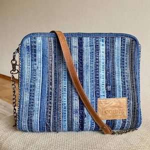 Könnte beinhalten: Eine blaue Jeans-Umhängetasche mit einem braunen Lederriemen und einem Metallkettenriemen. Die Tasche hat einen Aufnäher mit der Aufschrift "Original by Collins Jeanswear".
