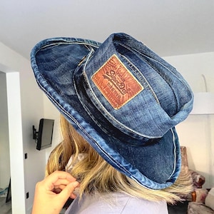 Puede incluir: Un sombrero de vaquero de mezclilla azul con un parche de cuero marrón con el texto "Superdry". El sombrero está hecho de tela de mezclilla y tiene un ala ancha. Un accesorio de moda.