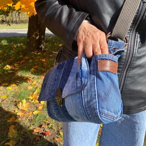 K&ouml;nnte beinhalten: Jeans-Umh&auml;ngetasche mit Klappenverschluss und braunem Lederaufn&auml;her. Die Tasche hat einen grauen Riemen und besteht aus recyceltem Jeansstoff. Die Tasche wird von einer Person getragen, die eine schwarze Lederjacke tr&auml;gt.
