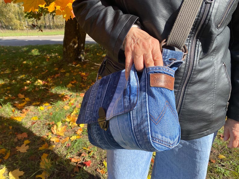 K&ouml;nnte beinhalten: Jeans-Umh&auml;ngetasche mit Klappenverschluss und braunem Lederaufn&auml;her. Die Tasche hat einen grauen Riemen und besteht aus recyceltem Jeansstoff. Die Tasche wird von einer Person getragen, die eine schwarze Lederjacke tr&auml;gt.
