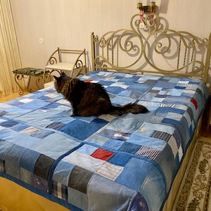 以下が含まれることがあります： 金色の金属製のヘッドボードが付いたベッドの上に、青いデニムのキルトパッチワークの上に、茶色のトラ猫が座っています。 キルトは、さまざまな色の青いデニムの正方形でできており、中央に赤い正方形があります。