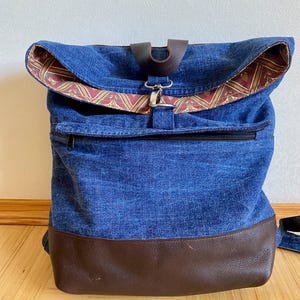 Könnte beinhalten: Ein blauer Jeans-Rucksack mit braunen Lederakzenten. Der Rucksack hat einen Klappverschluss und eine Reißverschlusstasche auf der Vorderseite.