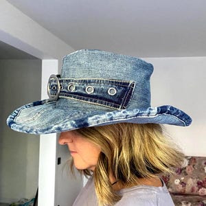 May include: A denim cowboy hat with a wide brim and a structured crown. The hat features a silver-toned buckle and grommet details. The hat is made of light blue denim.