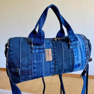 May include: A blue denim duffel bag with a zipper closure, top handles, and a detachable shoulder strap. The bag is made from repurposed denim and features a patchwork design with various shades of blue. A small rectangular tag is sewn on the side.
