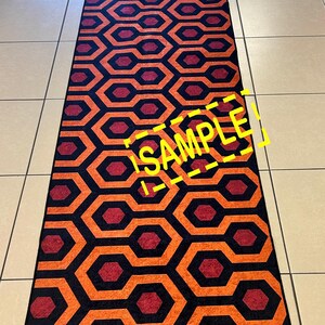 Op de afbeelding: Een lang, rechthoekig tapijt met een herhalend geometrisch patroon. Het ontwerp bevat oranje, rode en zwarte in elkaar grijpende vormen. Het woord "SAMPLE" is in het geel over het tapijt zichtbaar.