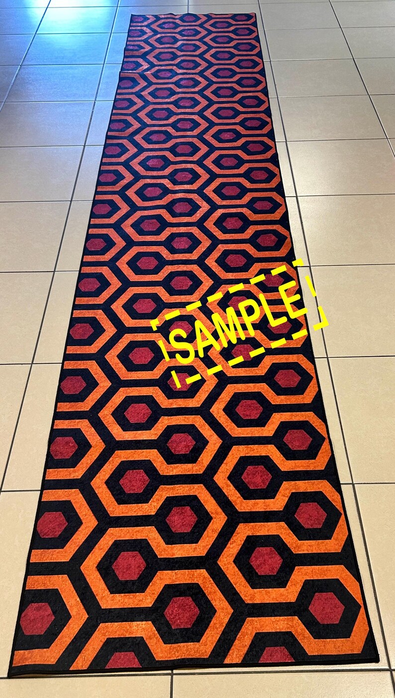 Op de afbeelding: Een lang, rechthoekig tapijt met een herhalend geometrisch patroon. Het ontwerp bevat oranje, rode en zwarte in elkaar grijpende vormen. Het woord "SAMPLE" is in het geel over het tapijt zichtbaar.