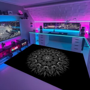 May include: A modern gaming room with a large screen displaying a video game. The room features a black rug with a white mandala design, a desk with multiple monitors, and neon lighting in shades of pink and blue. The words "GAME OVER" are visible.