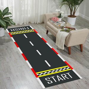 Op de afbeelding: Een rechthoekig tapijt dat is ontworpen om op een racebaan te lijken. Het heeft een zwart oppervlak met witte rijstrookmarkeringen, rood-witte randen en gele geblokte lijnen. De woorden "START" en "FINISH" staan aan elk uiteinde.