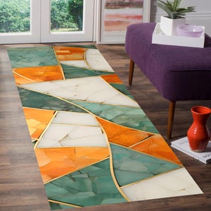 Op de afbeelding: Een rechthoekig tapijt met een geometrisch ontwerp. Het tapijt heeft een patroon van onregelmatige vormen in tinten oranje, blauwgroen en wit, omlijnd met gouden lijnen. Het tapijt ligt op een houten vloer, met een glimp van een deur en een paarse poef op de achtergrond.