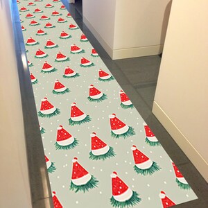 Op de afbeelding: Een lange, rechthoekige vloerkleed met een herhalend patroon van rode kerstmutsen op een lichtgrijze achtergrond. De mutsen hebben witte randen en zijn omgeven door groen, met kleine witte stippen verspreid.