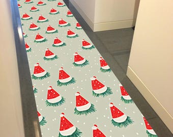 Op de afbeelding: Een lange, rechthoekige vloerkleed met een herhalend patroon van rode kerstmutsen op een lichtgrijze achtergrond. De mutsen hebben witte randen en zijn omgeven door groen, met kleine witte stippen verspreid.