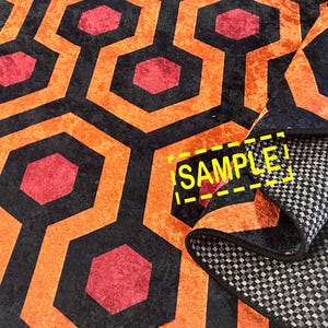 Op de afbeelding: Een patroonkleed met een herhalend geometrisch ontwerp. Het kleed heeft een zwarte achtergrond met oranje en rode zeshoekige vormen. Het kleed is opgevouwen en onthult een grijs en wit geblokt patroon aan de onderkant. Het woord "SAMPLE" is zichtbaar.
