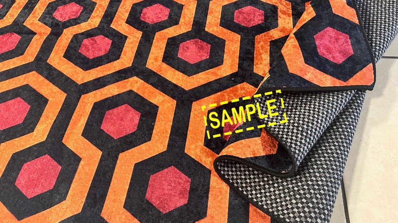 Op de afbeelding: Een patroonkleed met een herhalend geometrisch ontwerp. Het kleed heeft een zwarte achtergrond met oranje en rode zeshoekige vormen. Het kleed is opgevouwen en onthult een grijs en wit geblokt patroon aan de onderkant. Het woord "SAMPLE" is zichtbaar.