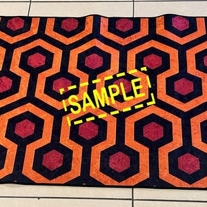 Op de afbeelding: Een rechthoekig tapijt met een herhalend geometrisch patroon van oranje, zwarte en rode zeshoeken. Het woord "SAMPLE" is in het geel op het tapijt gedrukt. Het tapijt ligt op een betegelde vloer.