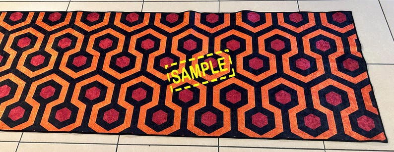 Op de afbeelding: Een rechthoekig tapijt met een herhalend geometrisch patroon van oranje, zwarte en rode zeshoeken. Het woord "SAMPLE" is in het geel op het tapijt gedrukt. Het tapijt ligt op een betegelde vloer.