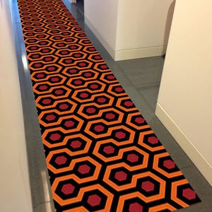 Op de afbeelding: Een lang, smal looptapijt met een herhalend geometrisch patroon van zwarte, oranje en rode zeshoeken. Het tapijt ligt in een gang.