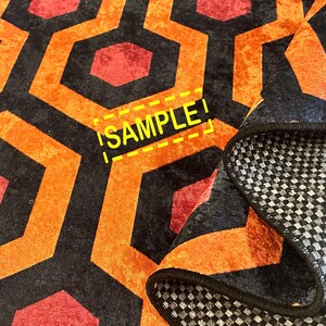 Op de afbeelding: Een opgevouwen tapijt met een herhalend geometrisch patroon van oranje, zwarte en rode zeshoeken. De onderkant van het tapijt is zichtbaar, met een grijs en wit geblokt ontwerp. Het woord "SAMPLE" is zichtbaar.