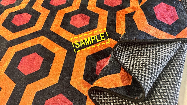 Op de afbeelding: Een opgevouwen tapijt met een herhalend geometrisch patroon van oranje, zwarte en rode zeshoeken. De onderkant van het tapijt is zichtbaar, met een grijs en wit geblokt ontwerp. Het woord "SAMPLE" is zichtbaar.