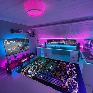 Può includere: Una sala giochi con una grande scrivania per computer, più monitor e una TV a schermo grande. La stanza è illuminata con luci al neon rosa e blu. La scrivania ha un grande tappetino per mouse con un design a circuito stampato. La TV mostra la parola "GHOST".