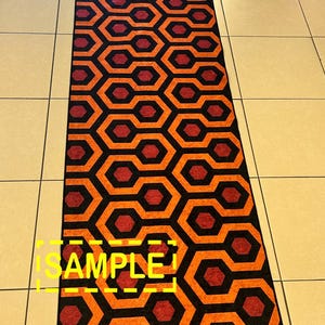 Op de afbeelding: Een lang, rechthoekig tapijt met een herhalend geometrisch patroon. Het ontwerp bevat zwarte, oranje en bordeauxrode vormen. Het woord "SAMPLE" is in het geel zichtbaar. Het tapijt ligt op een betegelde vloer.